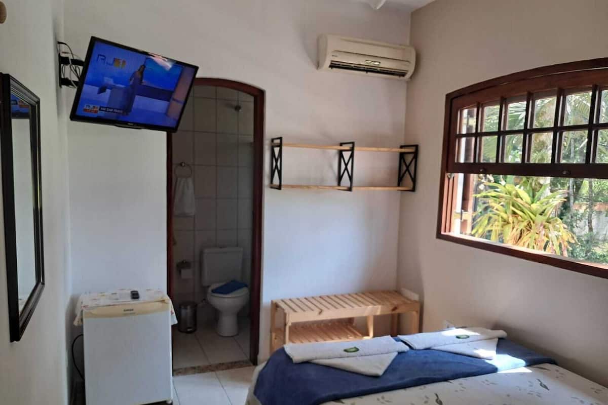 Quarto pequeno e limpo com uma cama, televisão, ar condicionado e janela com vista para áreas verdes.