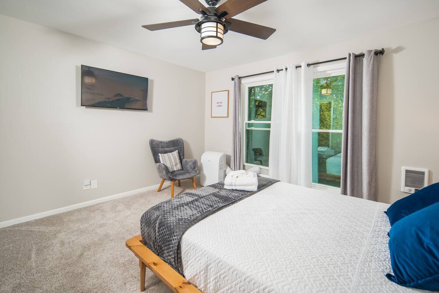 A bedroom with a bed, a chair, a ceiling fan, a window with curtains, and a painting on the wall.