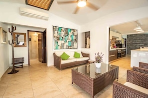 A modern living room with a sofa, coffee table, and wicker chairs. There is a kitchen visible in the background.