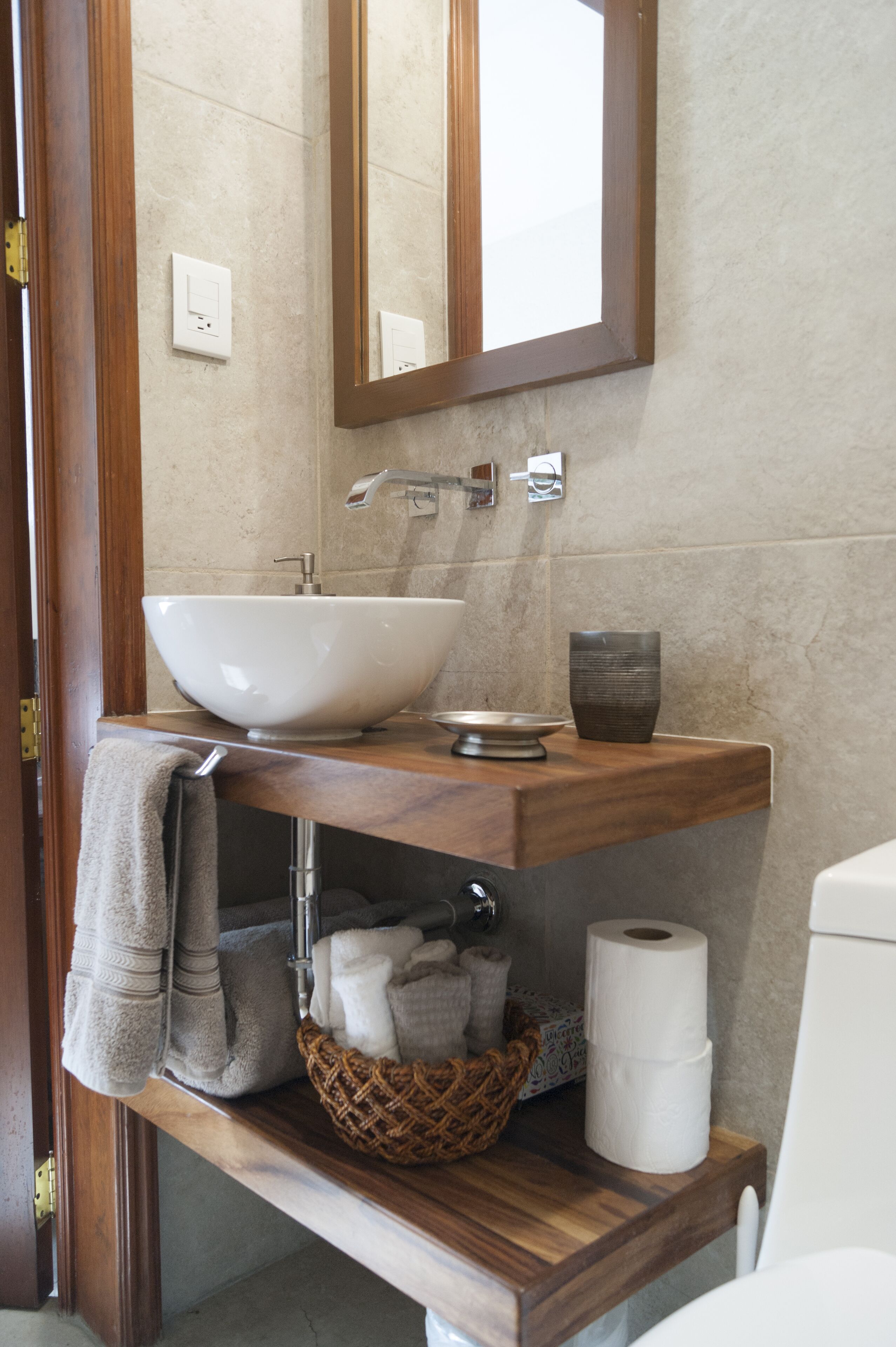 Un baño con tocador de madera, un lavabo blanco, un espejo, un perchero para toallas, una cesta y un dispensador de papel higiénico.