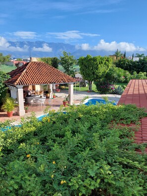 A covered outdoor seating area with a pool, surrounded by lush greenery and a mountainous backdrop.
