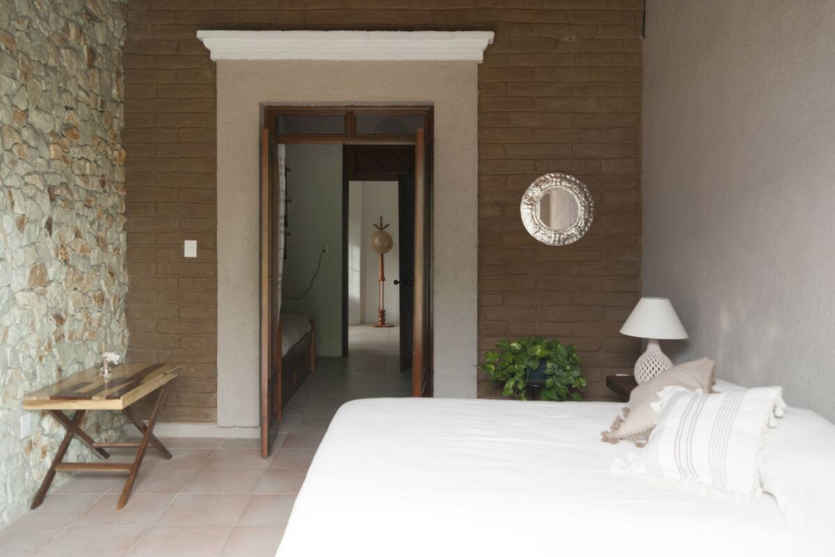 Un dormitorio con cama, mesita de noche, lámpara, espejo y planta en maceta.