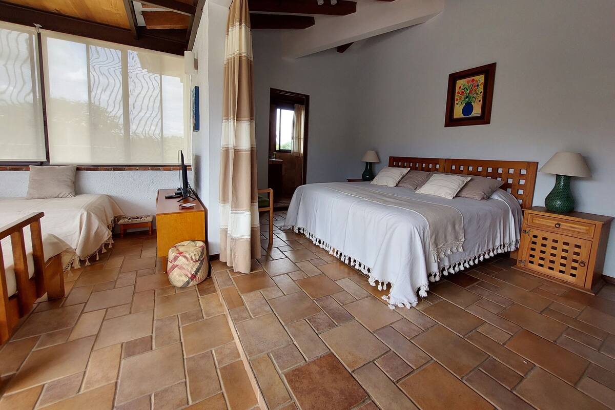 Un dormitorio con una cama grande, una cama más pequeña, un escritorio y una silla. Hay una ventana con cortinas, una lámpara y un cuadro en la pared.