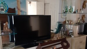 Ett vardagsrum med en tv på ett tv-bord, en byrå med lådor, en spegel och hyllor med dekorativa föremål.