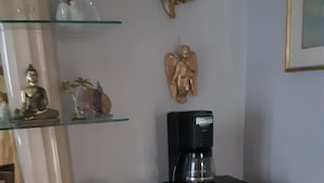 Ett rum med en beige byrå med lådor, en svart kaffebryggare på ett svart underställ, en glashylla med dekorativa föremål och en skulptur som hänger på väggen.
