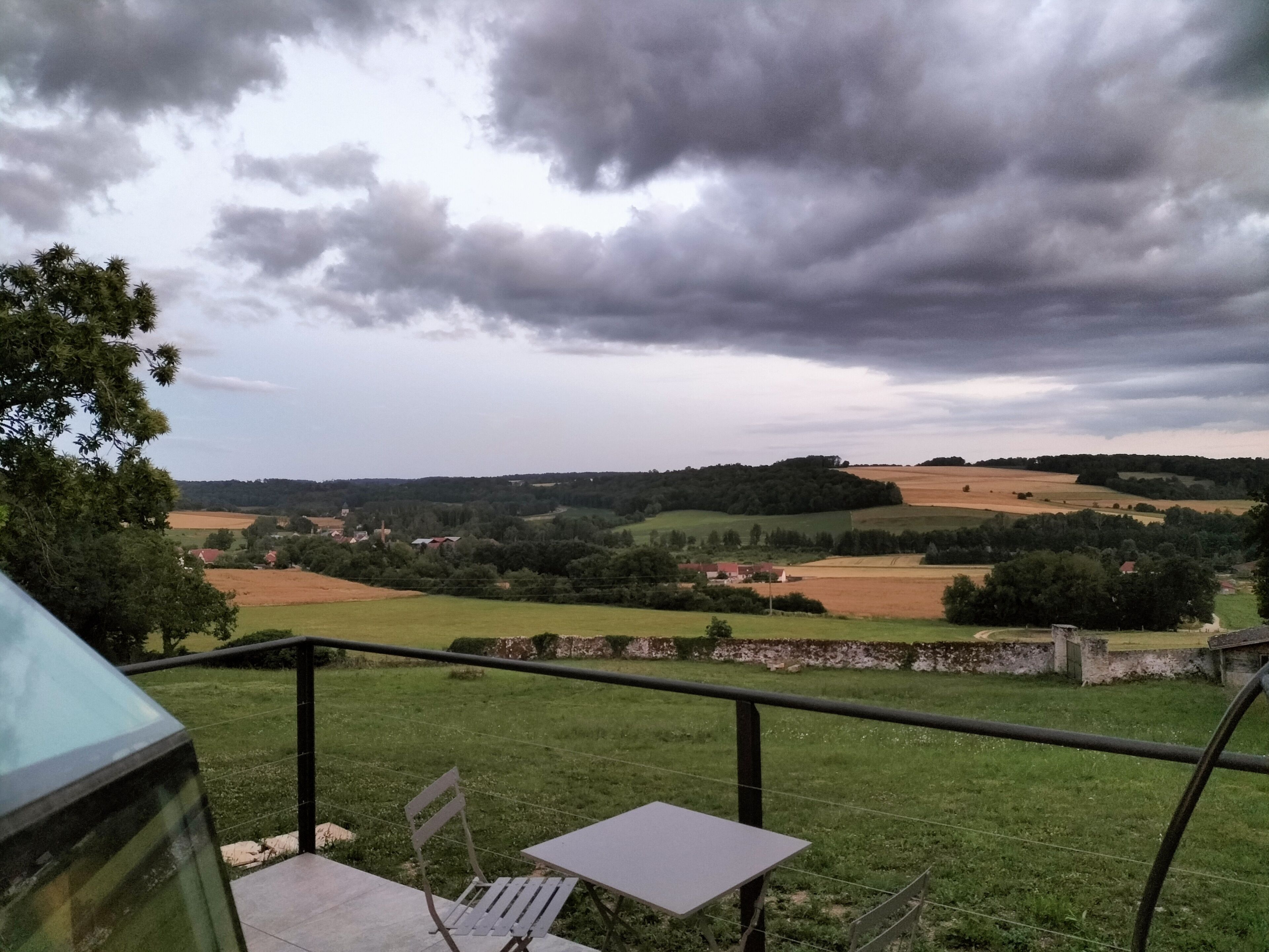 Una terraza con mesa y sillas, con vistas a un paisaje rural con campos y árboles bajo un cielo nublado.