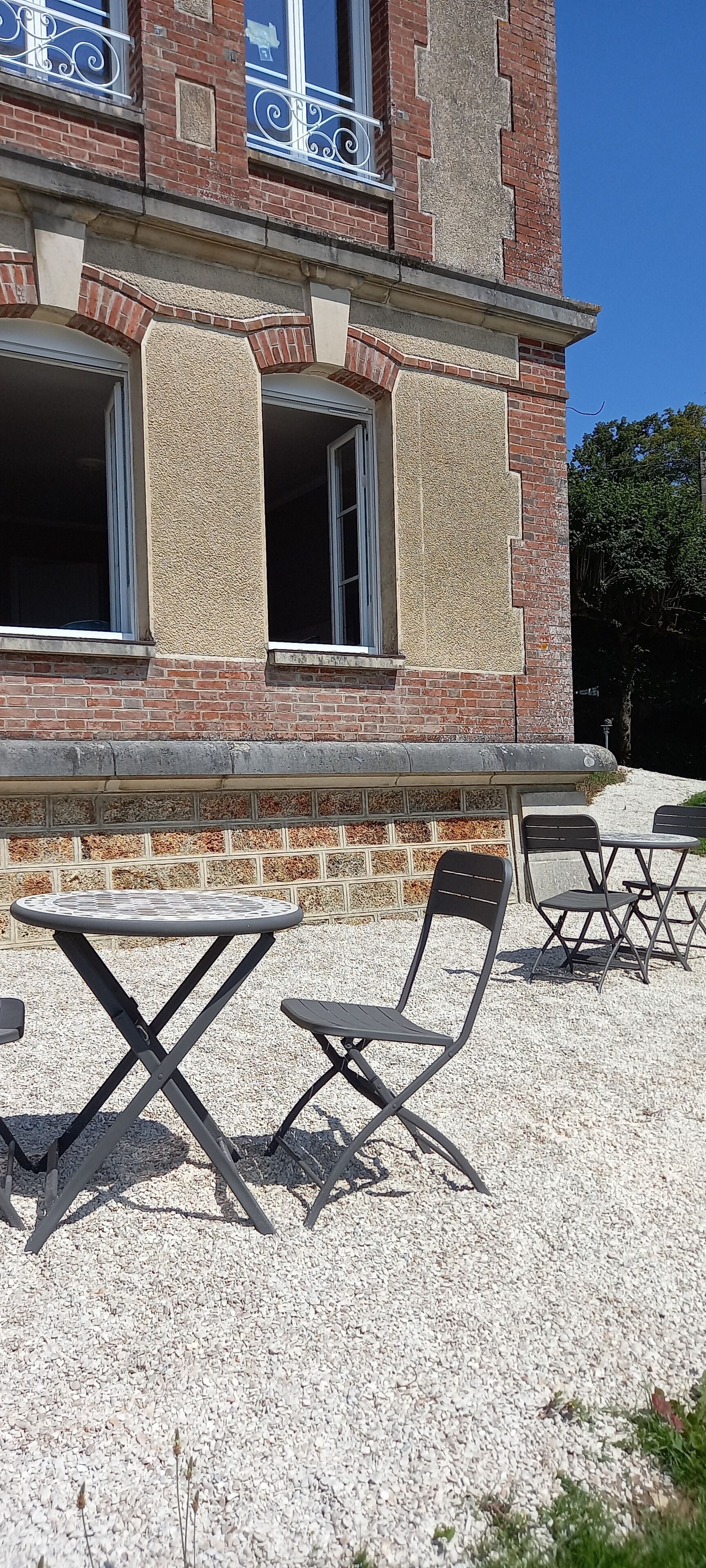 Un bâtiment à la façade en briques, doté de grandes fenêtres et d’un espace extérieur aménagé avec des tables et des chaises.