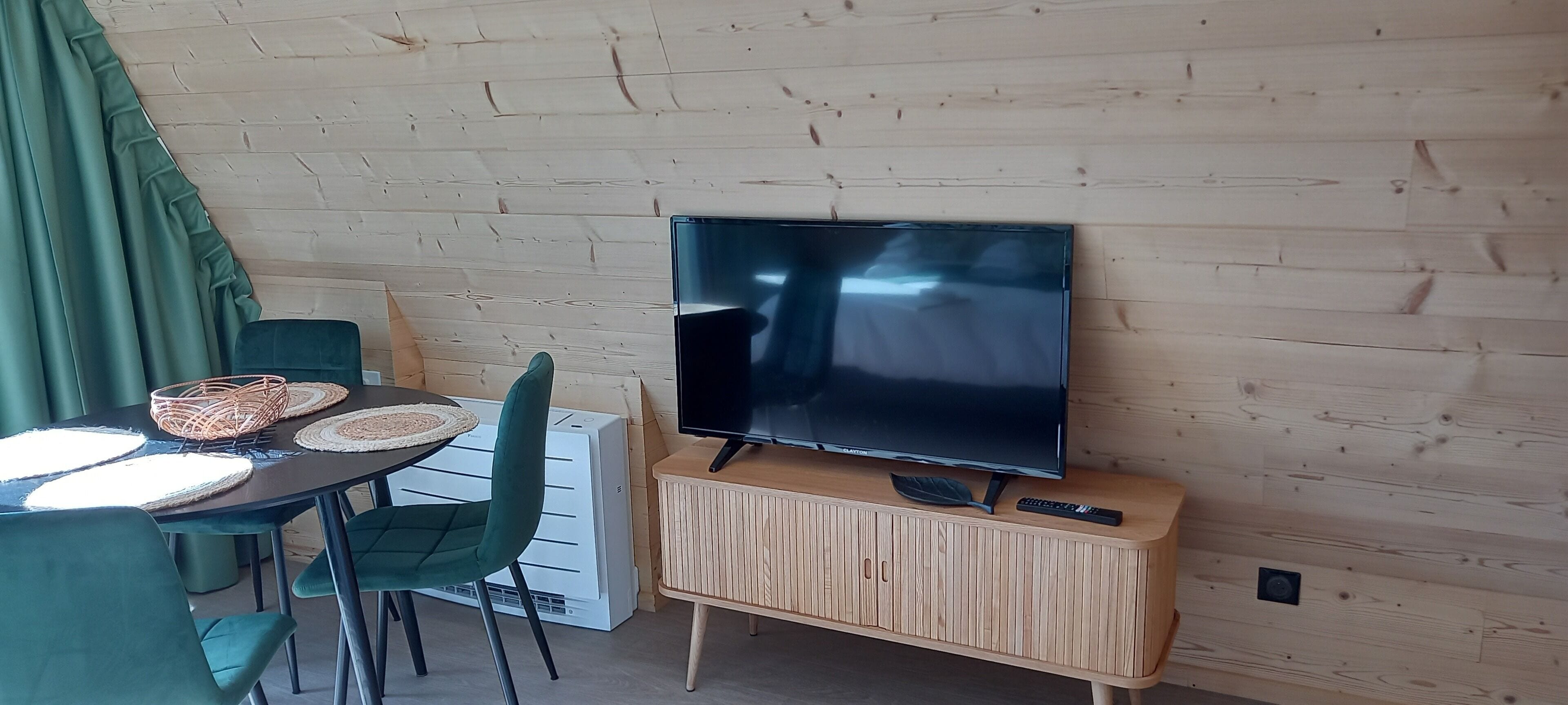 A room with a wooden wall, a flat-screen TV mounted on a wooden cabinet, a table with woven placemats, and green chairs.