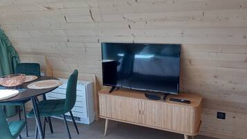 A room with a wooden wall, a flat-screen TV mounted on a wooden cabinet, a table with woven placemats, and green chairs.