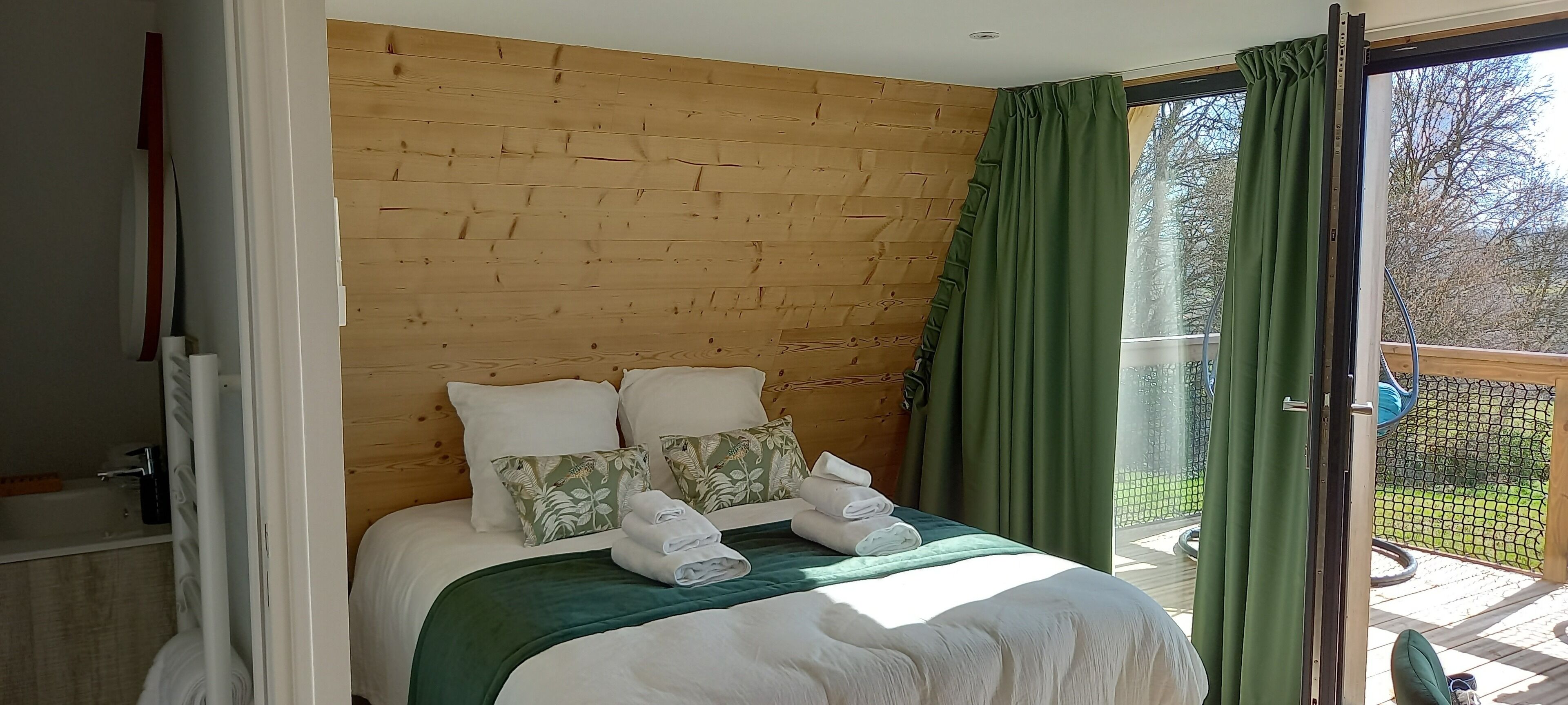 Quarto com cabeceira de madeira, cortina verde e vista para o exterior.