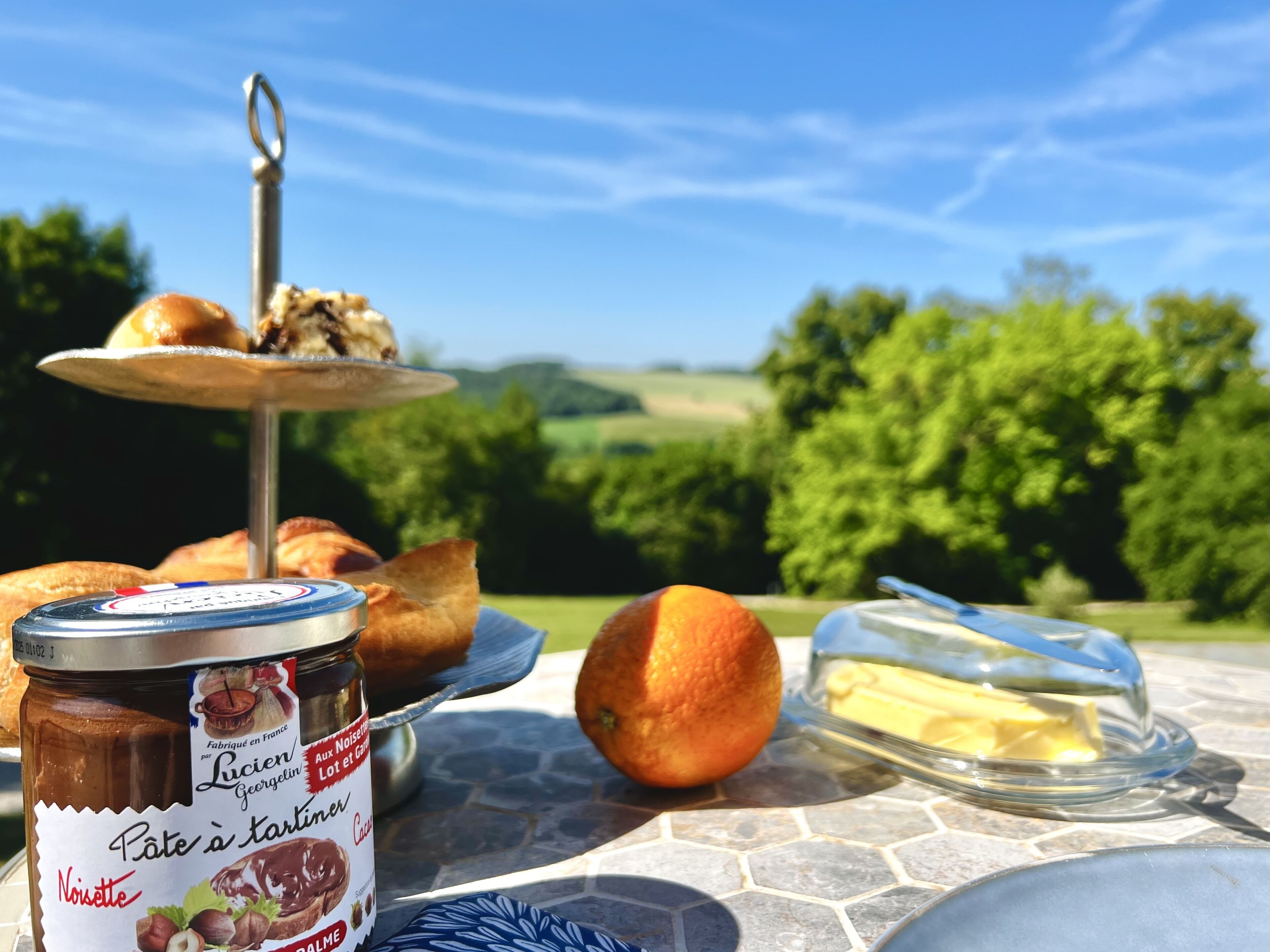 Ein Picknick-Arrangement mit einer Glaseinlage für Pastete, einem Teller Croissants, einer Orange und einer Butterpackung auf einem Steintisch im Freien.