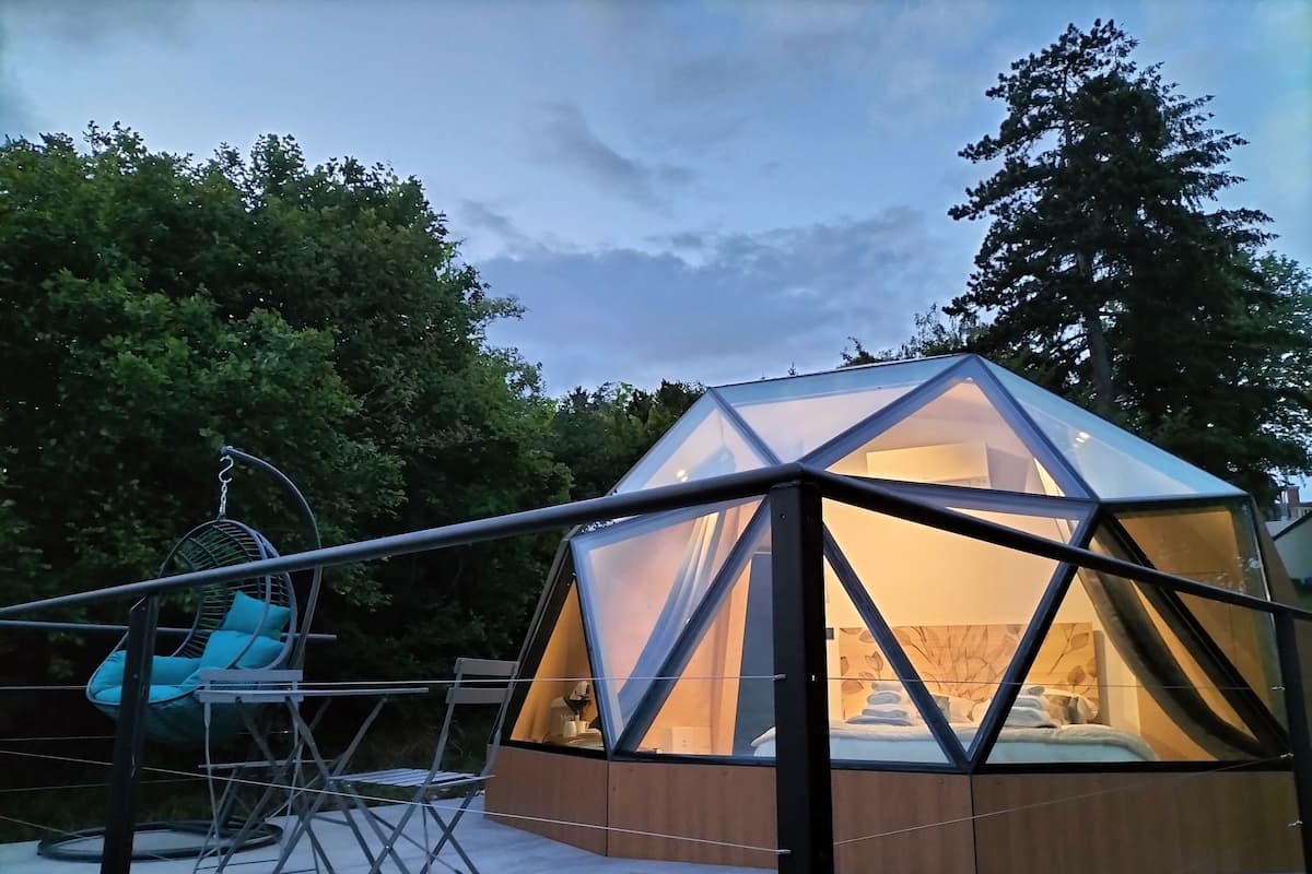 Une structure en forme de dôme géodésique abritant un lit, entourée d’une structure métallique et agrémentée d’un fauteuil suspendu.