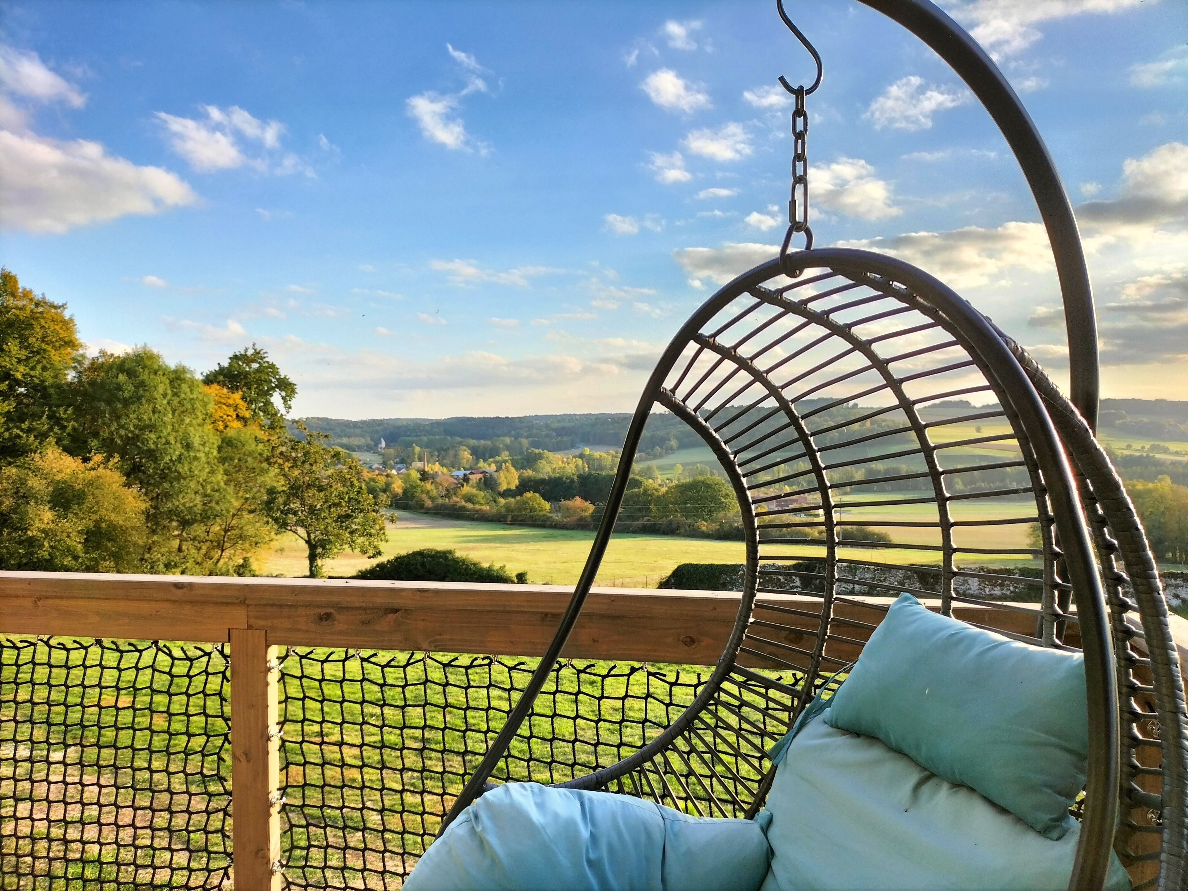 A wicker swing chair with cushions, overlooking a grassy landscape with trees and a distant view of hills.