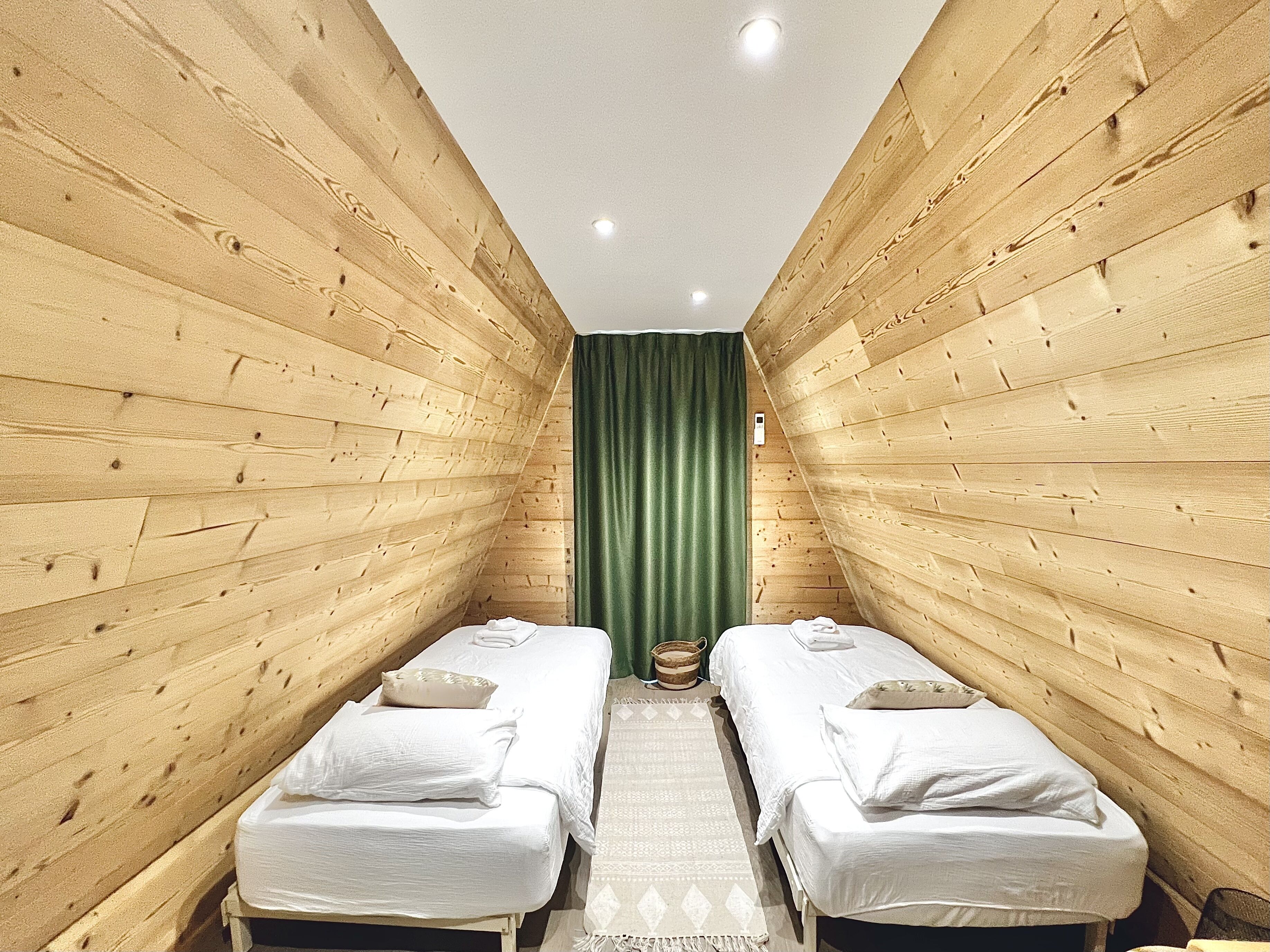 Une pièce étroite aux murs en bois, avec deux lits simples, un rideau vert et un petit tapis.