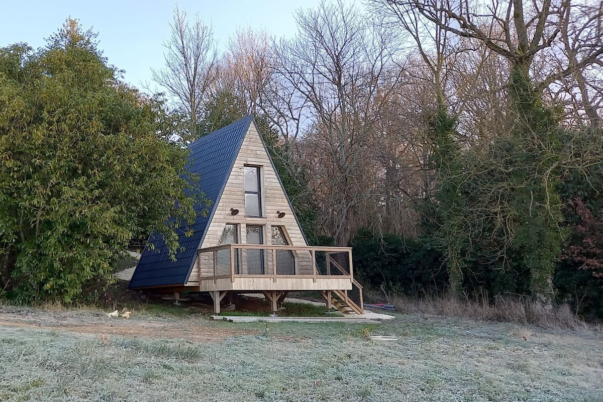 Une structure triangulaire en bois, dotée d’une fenêtre et d’une terrasse, entourée d’arbres et d’un sol recouvert de givre.