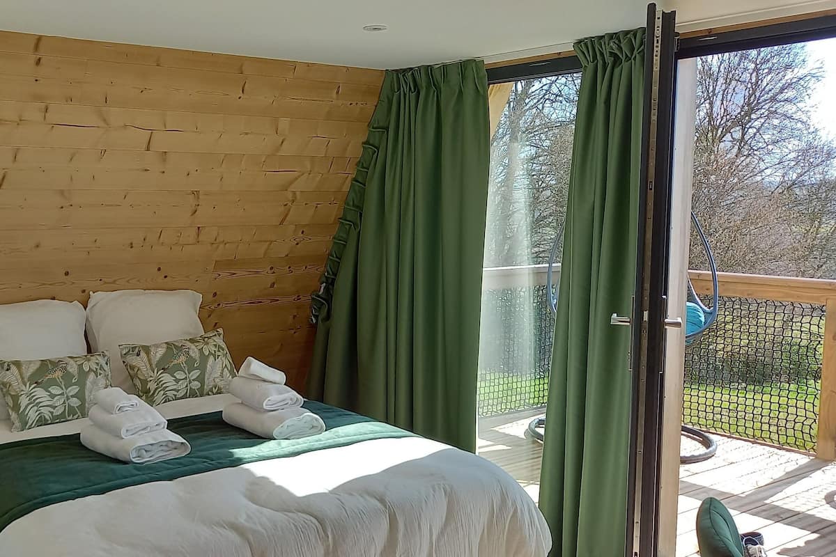 Une chambre avec une tête de lit en bois, un lit avec une literie blanche, des rideaux verts et une vue sur une terrasse.
