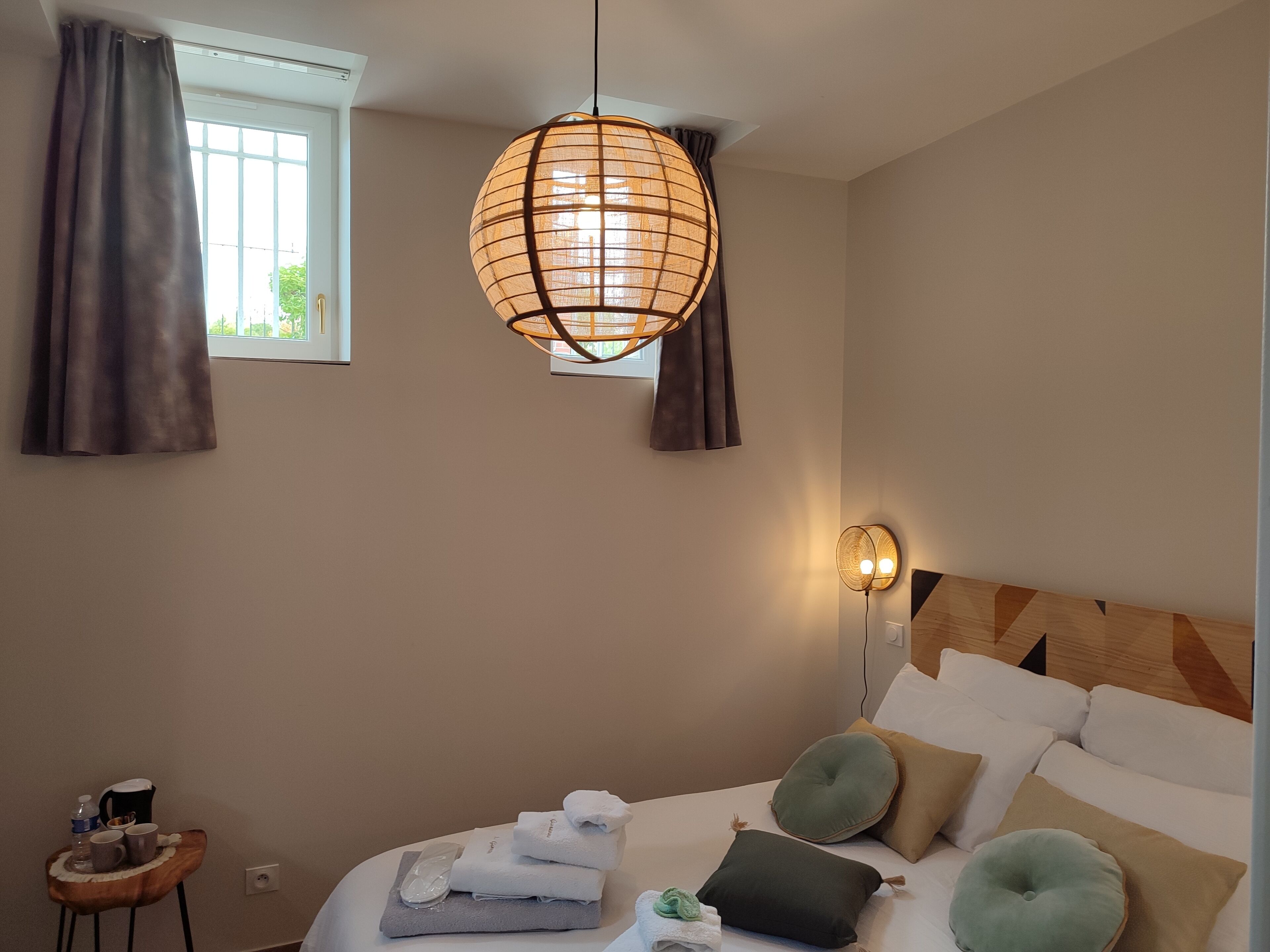 A hotel room with a bed, a bedside table with toiletries, a hanging lamp, and a window with curtains.