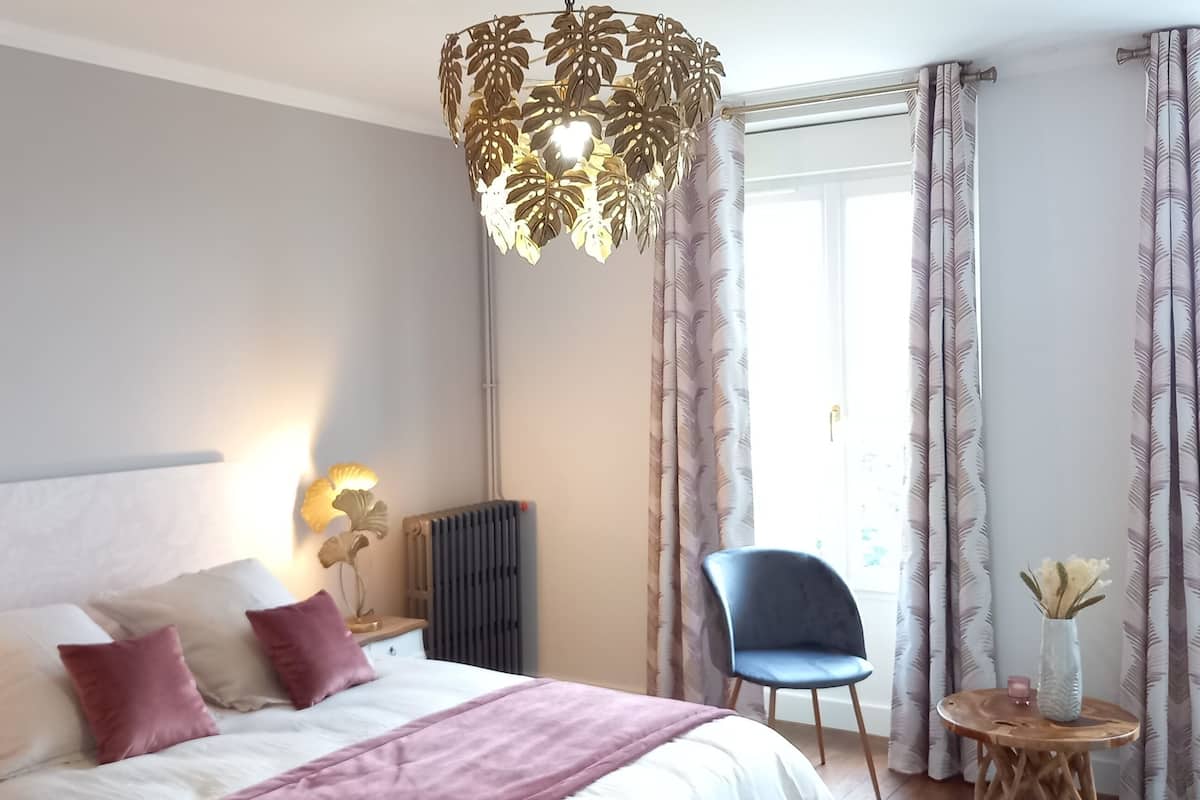 Une chambre à coucher avec un lit, des lampes de chevet, une chaise, une table et une fenêtre avec des rideaux.