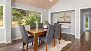 A dining area with a wooden table and grey upholstered chairs, large windows with a view of greenery, and a white console table with a vase of flowers. - Seaside Serenity - Kerikeri Holiday Home (Kerikeri)