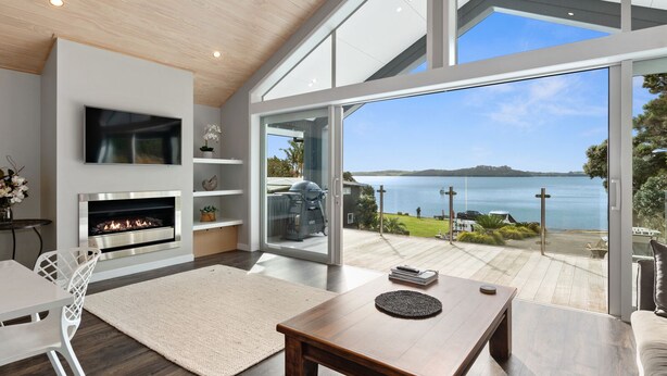 A modern living room with a fireplace, a TV, and a large glass door leading to a balcony with a view of the sea. - Seaside Serenity - Kerikeri Holiday Home (Kerikeri)