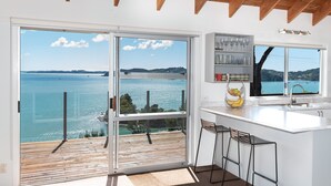 A modern kitchen with a large window overlooking a body of water, wooden deck, and bar stools. - Nelson's Lookout - Russell Holiday Home (Russell)