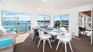 A modern dining area with a glass table, white chairs, and a view of the sea. - Nelson's Lookout - Russell Holiday Home (Russell)