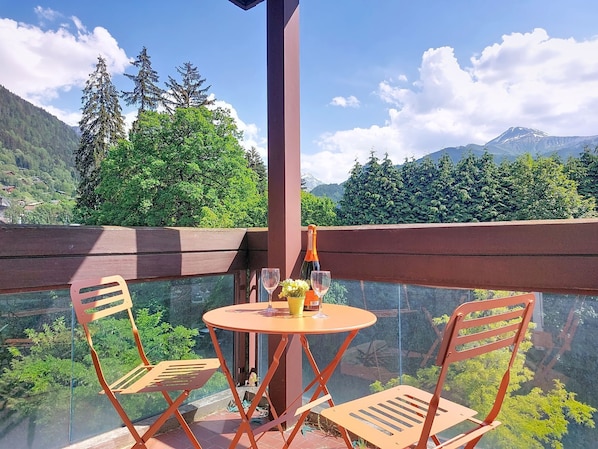 A balcony with a glass table and chairs, overlooking a mountainous landscape with trees and a clear sky. - Castel des roches A36 by Interhome (Saint Gervais)