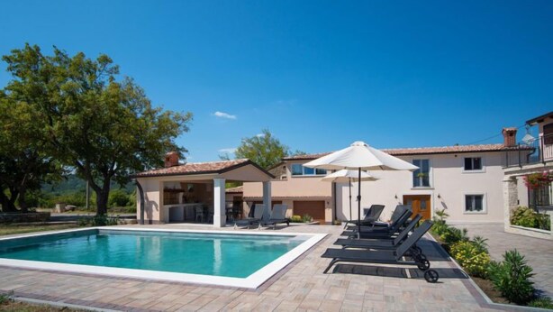 Piscina com espreguiçadeiras e guarda-sóis, rodeada por um jardim ajardinado e um edifício com telhado telhado.