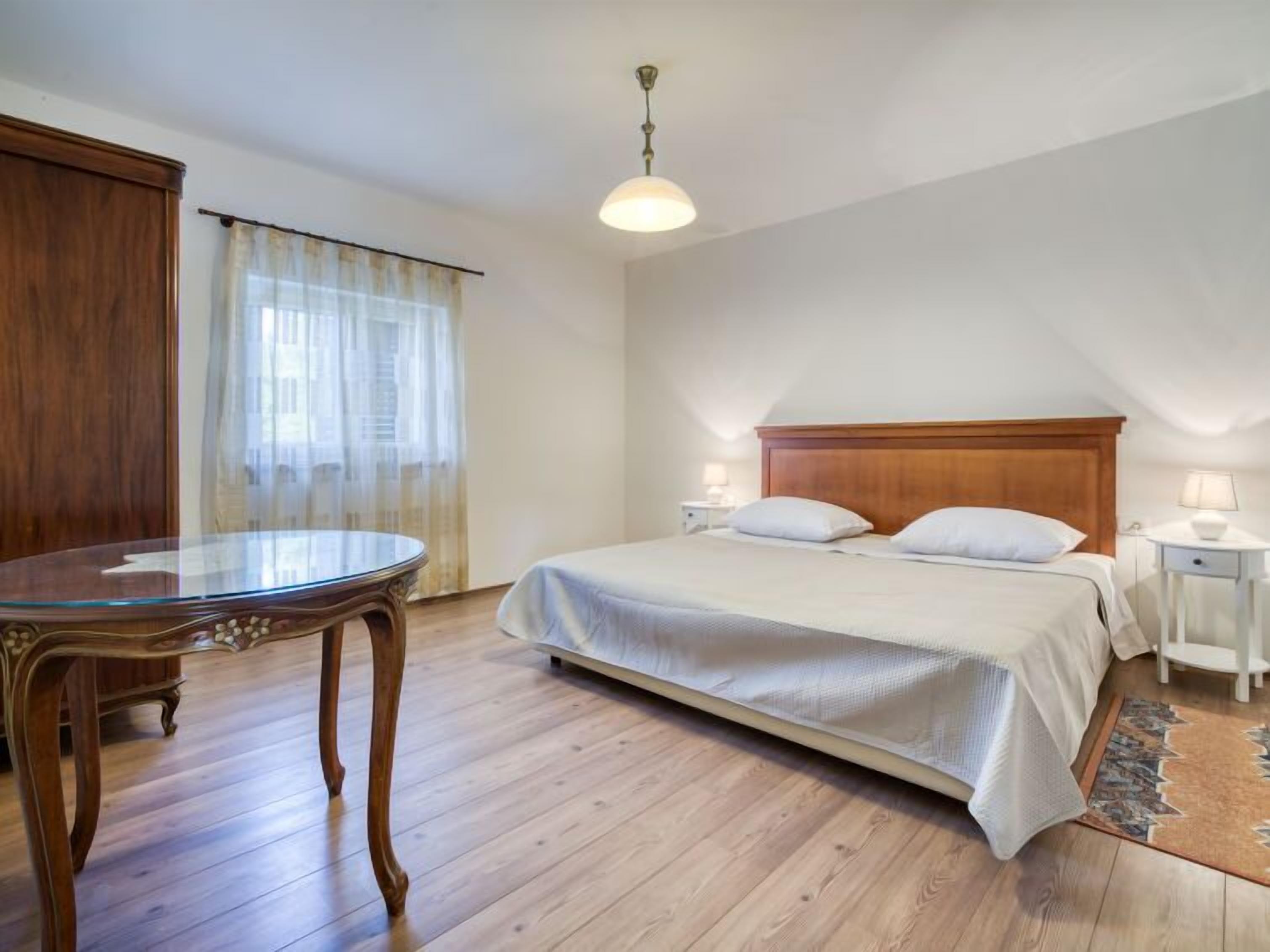 Une chambre à coucher avec un lit en bois, une table ronde, une fenêtre avec des rideaux et un luminaire suspendu.