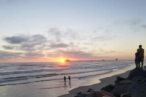 A beach scene with a sunset, rocky shoreline, and a pier in the background.