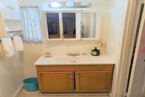 A bathroom with a white sink, wooden cabinets, a mirror, and a blue trash bin. - Cheerful,quiet, 2bd 1 bath home + patio, pool, , walk to beach (Oceanside)