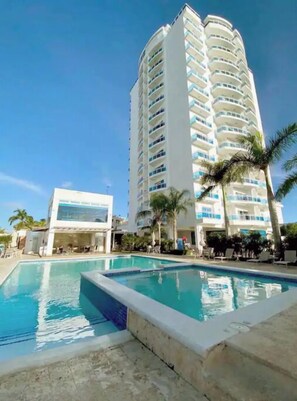 Um hotel com uma grande área de piscina, um edifício branco com várias varandas e palmeiras.