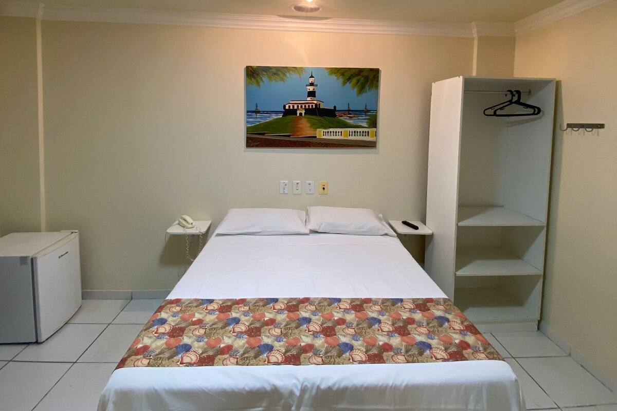 A hotel room with a bed, a painting of a lighthouse, a wardrobe, a desk with a phone, and a small refrigerator.