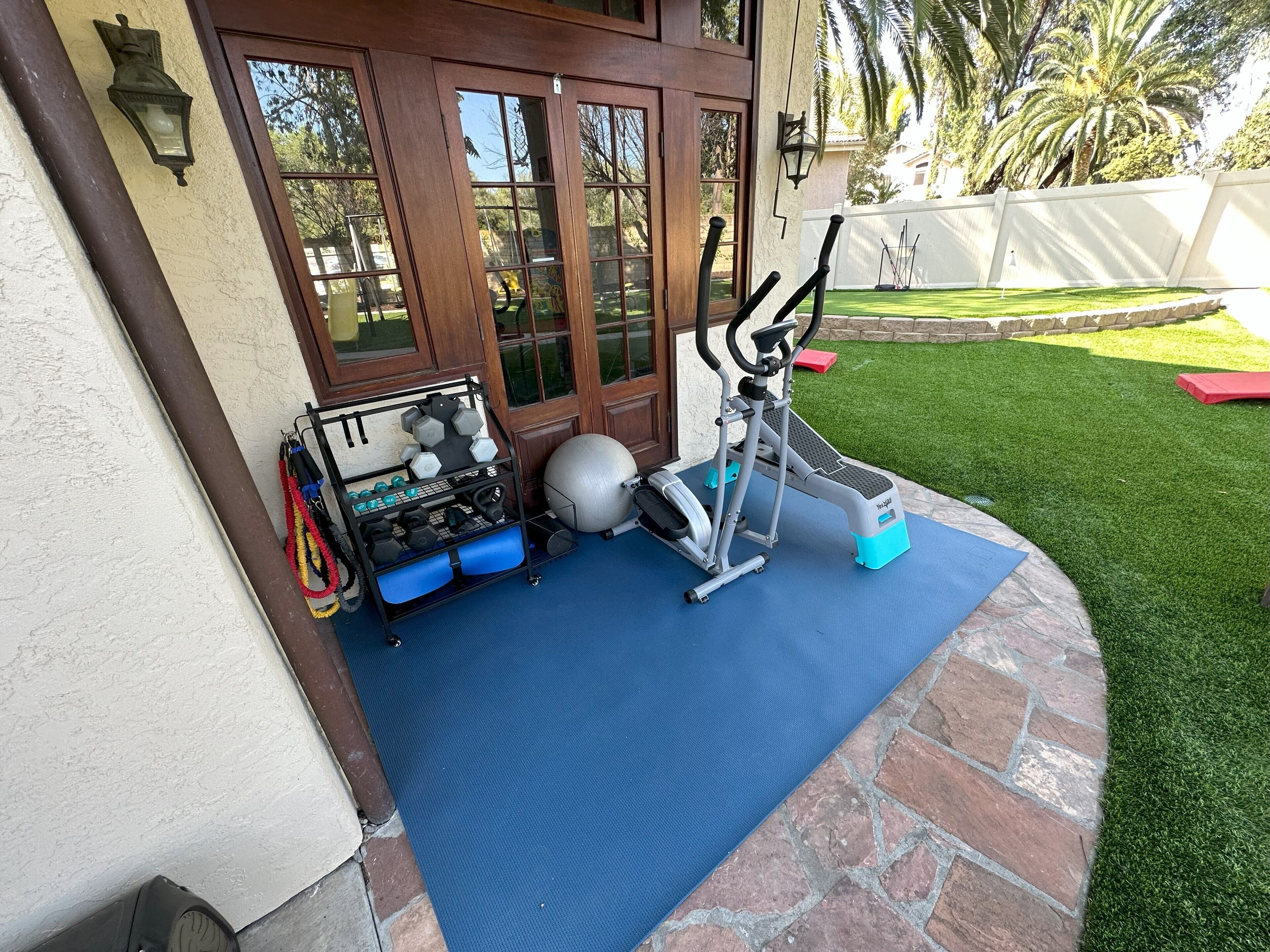 A home entrance with a blue mat, an exercise machine, and a rack with dumbbells and resistance bands.
