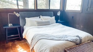 Ein Schlafzimmer mit einem Bett, einem Nachttisch, einer Lampe und Blick auf Bäume durch das Fenster.
