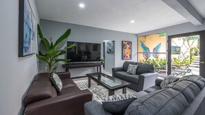 Ein modernes Wohnzimmer mit einem Flachbildfernseher, grauen Sofas, einem Couchtisch und großen Fenstern mit Blick ins Freie.