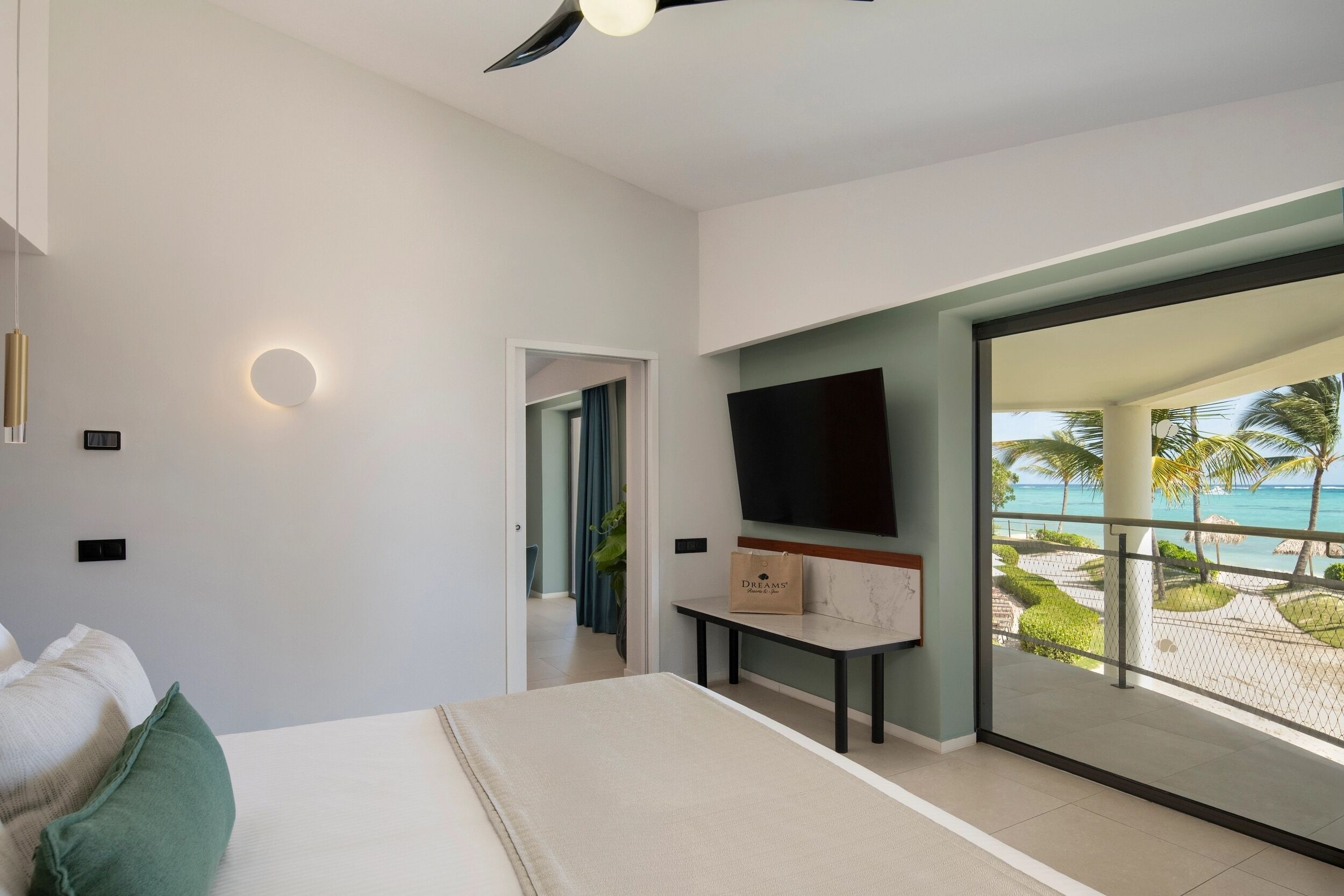 A modern hotel room with a bed, a TV, a desk, and a balcony with a view of the beach.