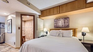 Quarto de hotel com uma cama grande, duas mesas de cabeceira com luminárias, uma cabeceira de madeira, um quadro na parede e uma porta que dá para um banheiro.