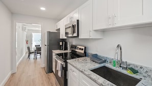 A modern kitchen with white cabinets, a stainless steel refrigerator, a microwave, a stove, and a sink with a dish rack. - Safari Apartments (Ocean City)