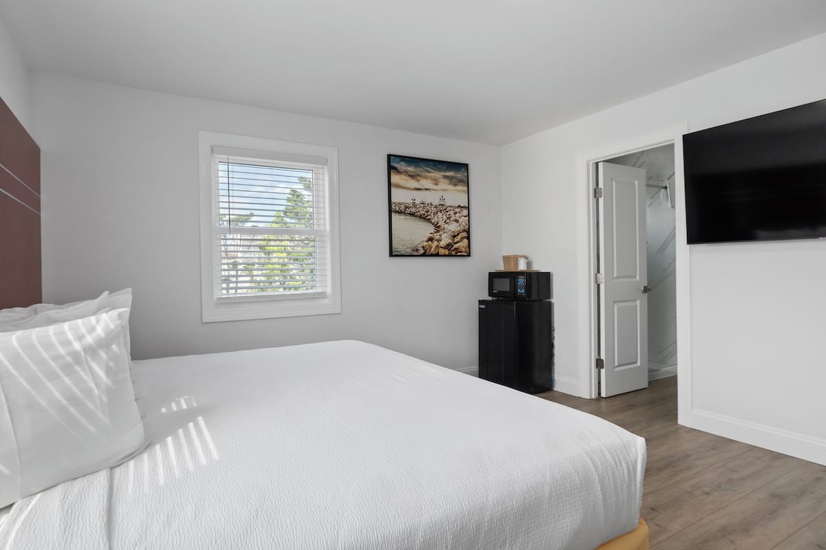 Un dormitorio con una cama, una ventana con persianas, un refrigerador, un cuadro y una puerta que da a otra habitación.