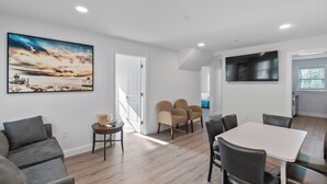A modern living room with a large painting, a dining area with a white table, and a kitchen visible through an open door. - Safari Apartments (Ocean City)