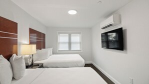 A hotel room with two beds, a TV mounted on the wall, a window with blinds, and an air conditioning unit. - Safari Apartments (Ocean City)
