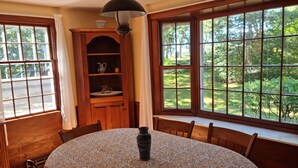 Un comedor con una mesa redonda, sillas, un armario de madera y ventanas amplias con vistas a la vegetación.