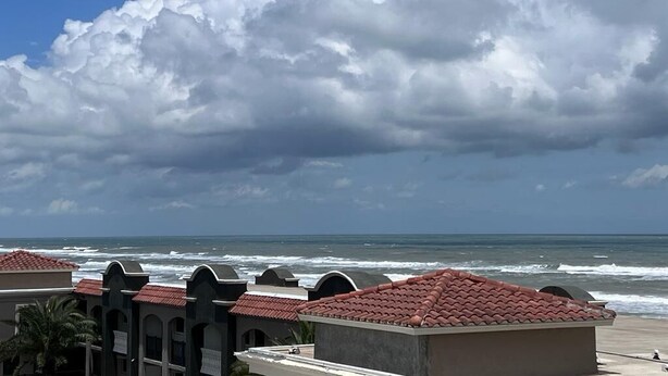 Un hotel frente al mar con techos de tejas rojas, vistas a la playa y un cielo grande cubierto de nubes.