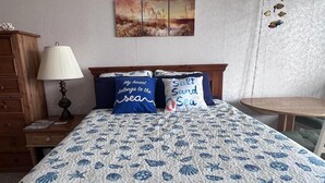 Un dormitorio con una cama con cubrecama azul y blanco con temática náutica, dos almohadas con texto, una mesita de noche de madera con una lámpara, un aparador de madera, y una pared con un cuadro y decoración.