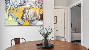 Une table à manger avec des chaises, un vase décoré de verdure et un tableau représentant un cheval et une roue de charrette accroché au mur.