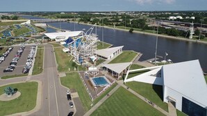 An aerial view of a waterfront property with a large parking lot, a swimming pool, and a complex of buildings with a distinctive white and blue architectural design.