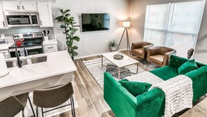 A modern kitchen with white cabinets and stainless steel appliances, a dining area with a white table and green upholstered chairs, and a living room with a green sofa and a floor lamp.