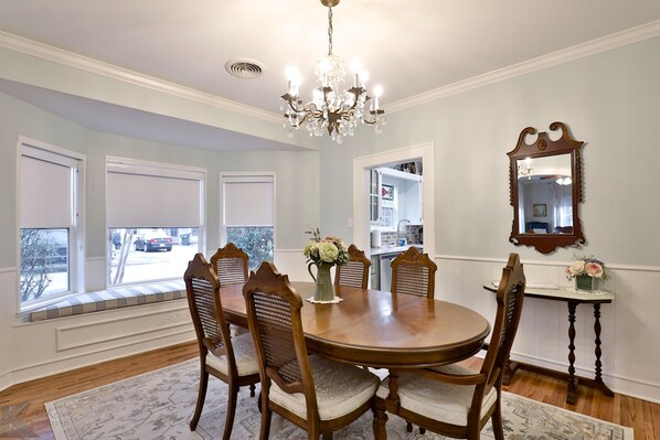 A dining room with a round wooden table, six chairs, a chandelier, a mirror, and a window seat.
