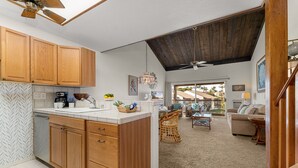A kitchen with wooden cabinets, a white countertop, and a ceiling fan. There is a dining area with a table and chairs, and a view of the outdoors. - Cozy Beachside Retreat Steps from Kamaole III (Kihei)